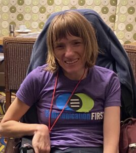 Melissa Crisp-Cooper smiling, sitting in a wheelchair, wearing a purple t-shirt with the CommunicationFIRST logo.