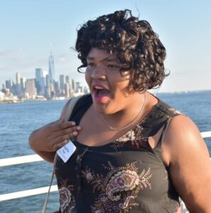 Angelica Vega, a woman in her early 20s with brown skin and curly brown hair wearing a sleeveless brown dress standing in front of a white boat railing with blue skies and a body of water in the background and Manhattan skyscrapers in the distance. She is wearing red lipstick and has her mouth open as if in pleasant surprise.