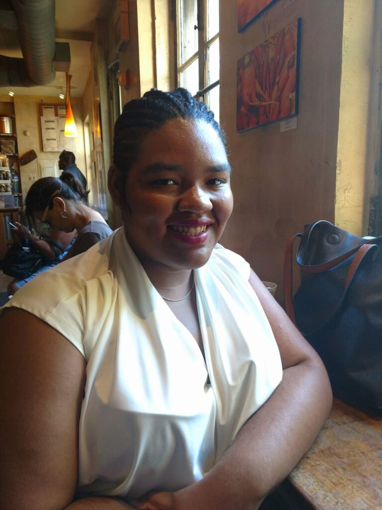 a young black woman with natural hair in cornrows sits smiling, looking at the camera, with her left elbow on a counter. Her face is lit by natural light from a window and she is earing a light top. In the background are people in the distance, apparently in a cafe or similar setting