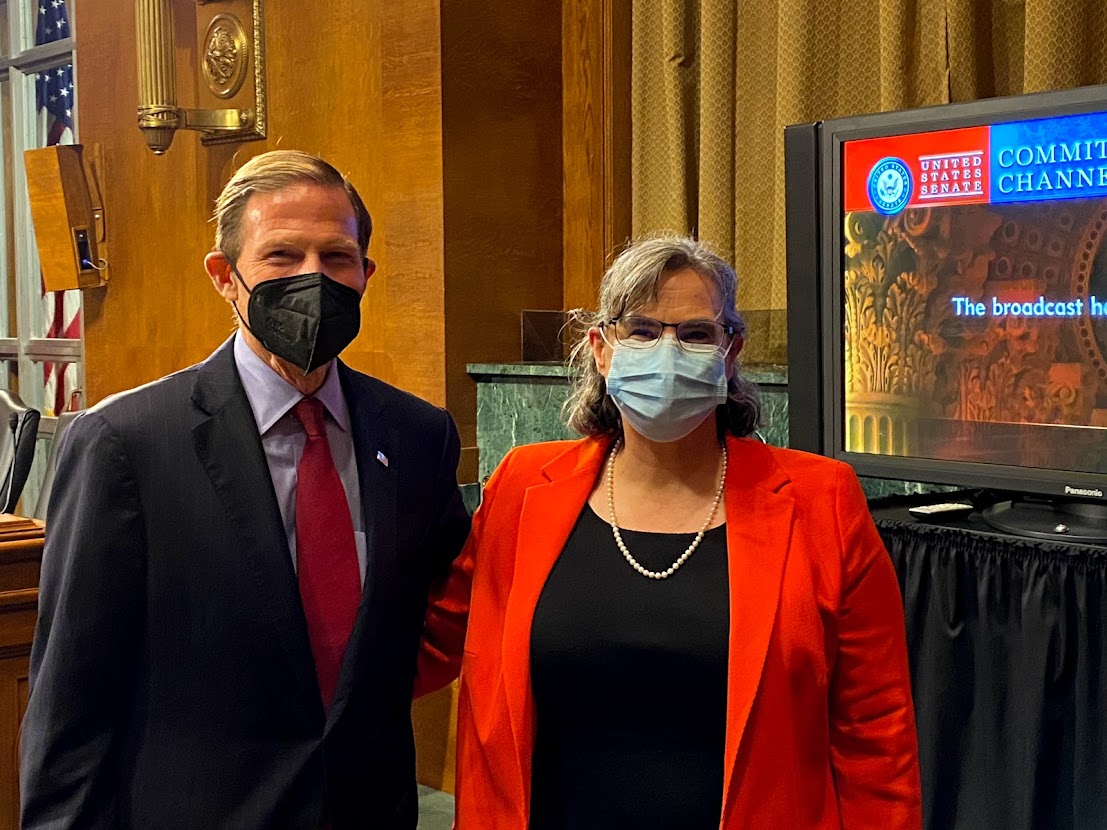 Dr. Clarissa Kripke, wearing a bright red jacket over a black dress, stands next to Senator Richard Blumenthal (D-CT). They are wearing masks, smiling, and looking at the camera. The wood-paneled wall of the Senate Judiciary Subcommittee hearing room is behind them, as is a television screen.
