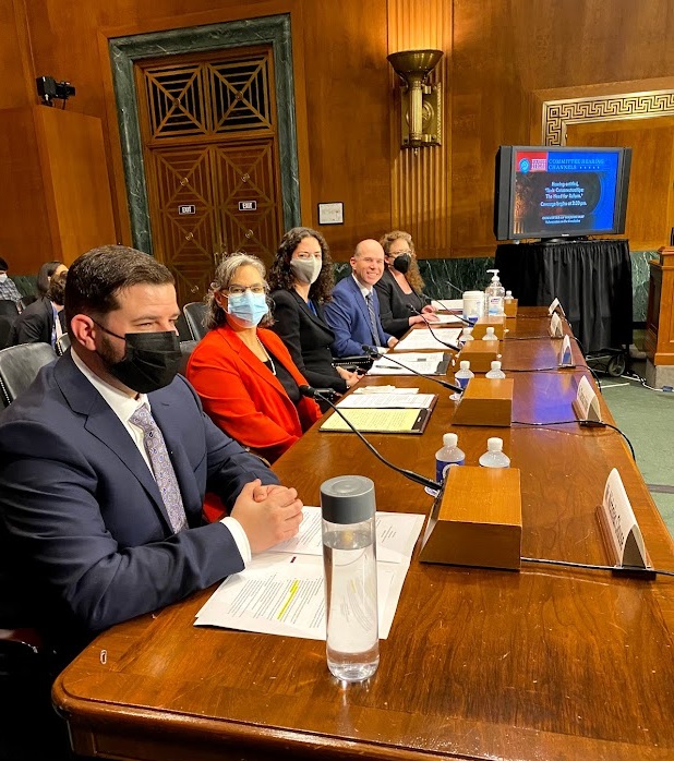 A photo of five witnesses wearing suits seated before a long wooden table with papers in front of them in a US Senate Judiciary Subcommittee hearing room. One of the witnesses is Dr. Clarissa Kripke, wearing a red jacket. Four of the five witnesses are wearing masks. Four of the five witnesses are looking at the camera and appear to be smiling underneath their masks.