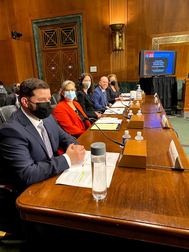 A photo of five witnesses wearing suits seated before a long wooden table with papers in front of them in a US Senate Judiciary Subcommittee hearing room. One of the witnesses is Dr. Clarissa Kripke, wearing a red jacket. Four of the five witnesses are wearing masks. Four of the five witnesses are looking at the camera and appear to be smiling underneath their masks.