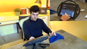 Young white man with brown hair sitting at a table typing on an iPad communication device. An inset of the screen he is typing on is visible. The screen and video captions read "Hi everyone."