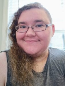 A nonbinary person with pale skin and wearing glasses smiles at the camera