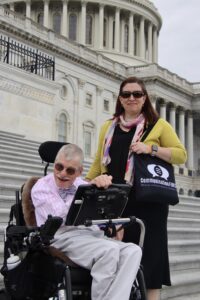 White woman with long dark hair holding a CommunicationFIRST bag and standing behind a white man with grey hair sitting in a power wheelchair with an AAC device. Both are wearing sunglasses and are smiling, with the US Capitol building behind them.