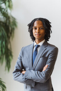 A Black man with dreadlocks wearing a suit and tie is looking at the camera with folded arms