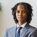A Black man with dreadlocks wearing a suit and tie is looking at the camera with folded arms