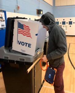 Ben Breaux, a smiling white man with facial hair, wearing a hoodie, noise-canceling headphones, and holding an AAC device, voting at an indoor polling place that appears to be a gym. A sign that says "Vote" with an American flag is obscuring the booth where he is standing and filling out his ballot.