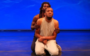 Two Black men kneeling on a stage facing the audience. One is behind the other and his hands are on the biceps of the man in front, who is wearing a grey tanktop and white trousers. Both men have serious looks on their faces. The man in back has long hair and a short beard, and his face is partially obscured by the man in front. The man in front is Lateef McLeod and he has very short hair with a hint of a beard. His hands are balled into relaxed fists and are laying on top of each other at the wrist. They are bathed in a warm light, and the background is bathed in an electric blue light.