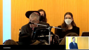 Bob Williams, an older white man sitting in a wheelchair and wearing a yarmulke looks at the audience while presenting in front of a microphone. A speech generating device is in front of him. Two women rabbis sit behind him looking on.