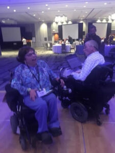 Bob Williams and Judy Heumann, sitting in their power wheelchairs, dance under purplish light on a conference dancefloor.