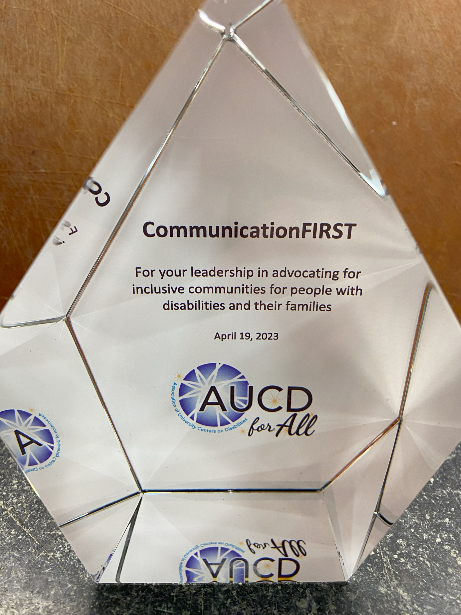 A head-on view of the AUCD award, which is a tall pentagon glass cube with the back of the cube lined in white with the text: "CommunicationFIRST - For your leadership in advocating for inclusive opportunities for people with disabilities and their families - April 19, 2023" the AUCD for All logo appears at the bottom.