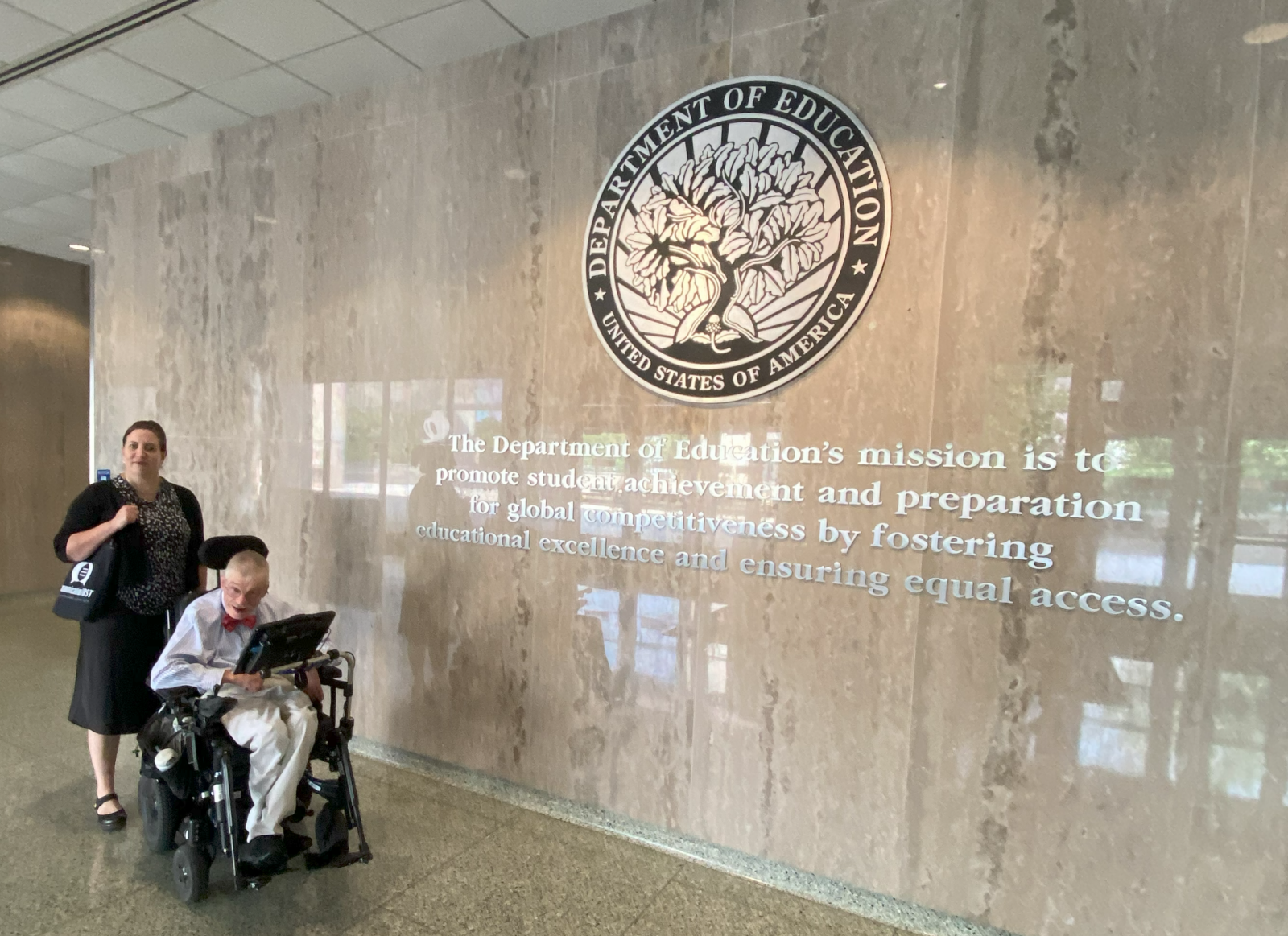CommunicationFIRST Executive Director Tauna Szymanski and Policy Director Bob Williams in front of the U.S. Department of Education (ED) seal. The seal is located in the lobby of the U.S. Department of Education building and hangs on a wall of marble. The words, “The Department of Education’s mission is to promote student achievement and preparation for global competitiveness by fostering educational excellence and ensuring equal access” are below the seal. Bob is a smiling white man with white hair and a mustache wearing glasses and a bowtie with a button-down shirt and khaki pants, sitting in a motorized wheelchair with an AAC device in front of him. Tauna is a smiling white woman with brown hair. She is standing behind Bob and is wearing black and holding a black bag with the CommunicationFIRST logo on it in white.