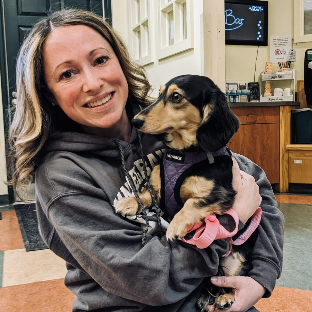 A smiling Jen Schonger, a white woman with long wavy hair, holding a dachshund in her arms at the vet.