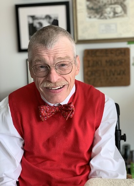 Headshot of Bob Williams, an older white man with white hair wearing a red vest and bowtie with a button up white-ish long-sleeved dress shirt. He is smiling at the camera in his home office, with various framed photos and a wooden letterboard behind him
