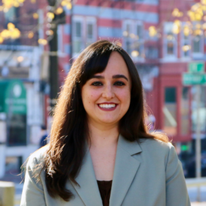 The subject of this headshot is Bre Mercier, a white woman in her thirties who is wearing her long, dark brown hair down and sporting a light green suit coat. On a sunny day, she smiles as she poses with city buildings and budding tree branches in the background behind her. 