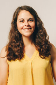 A woman with long, brown, wavy hair stands smiling at the camera. She is wearing a yellow sleeveless top.