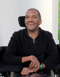 Lateef McLeod, a black man wearing a black fleece top sits in his power wheelchair smiling at the camera.