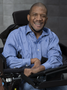 Lateef McLeod, a black man wearing a blue striped dress shirt, smiles at the camera from his power wheelchair.