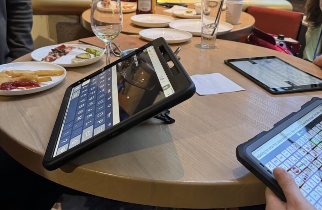 Three different speech generating devices are on a round table among drinking glasses and dinner plates. An AAC user's hand is in view as they type on one of the devices.