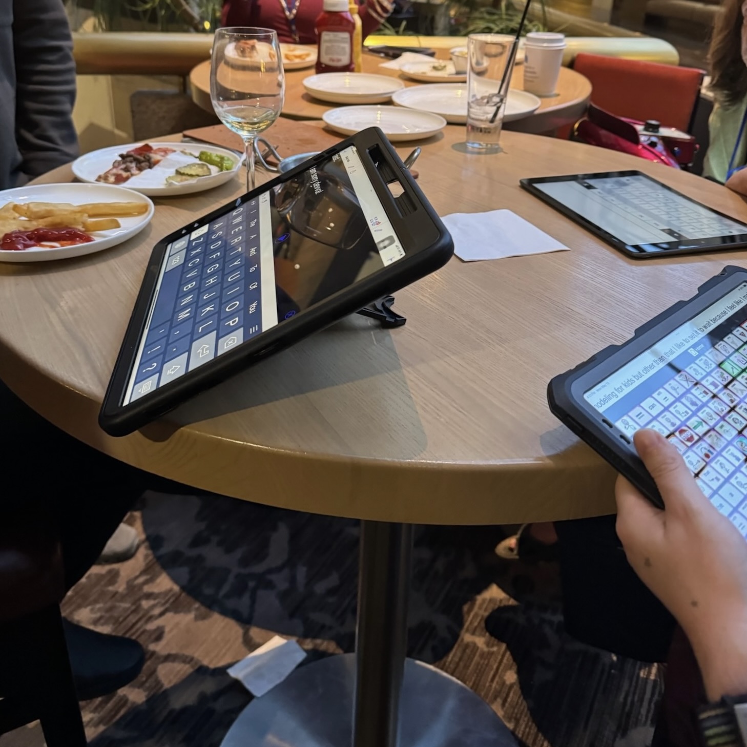 Three different speech generating devices are on a round table among drinking glasses and dinner plates. An AAC user's hand is in view as they type on one of the devices.