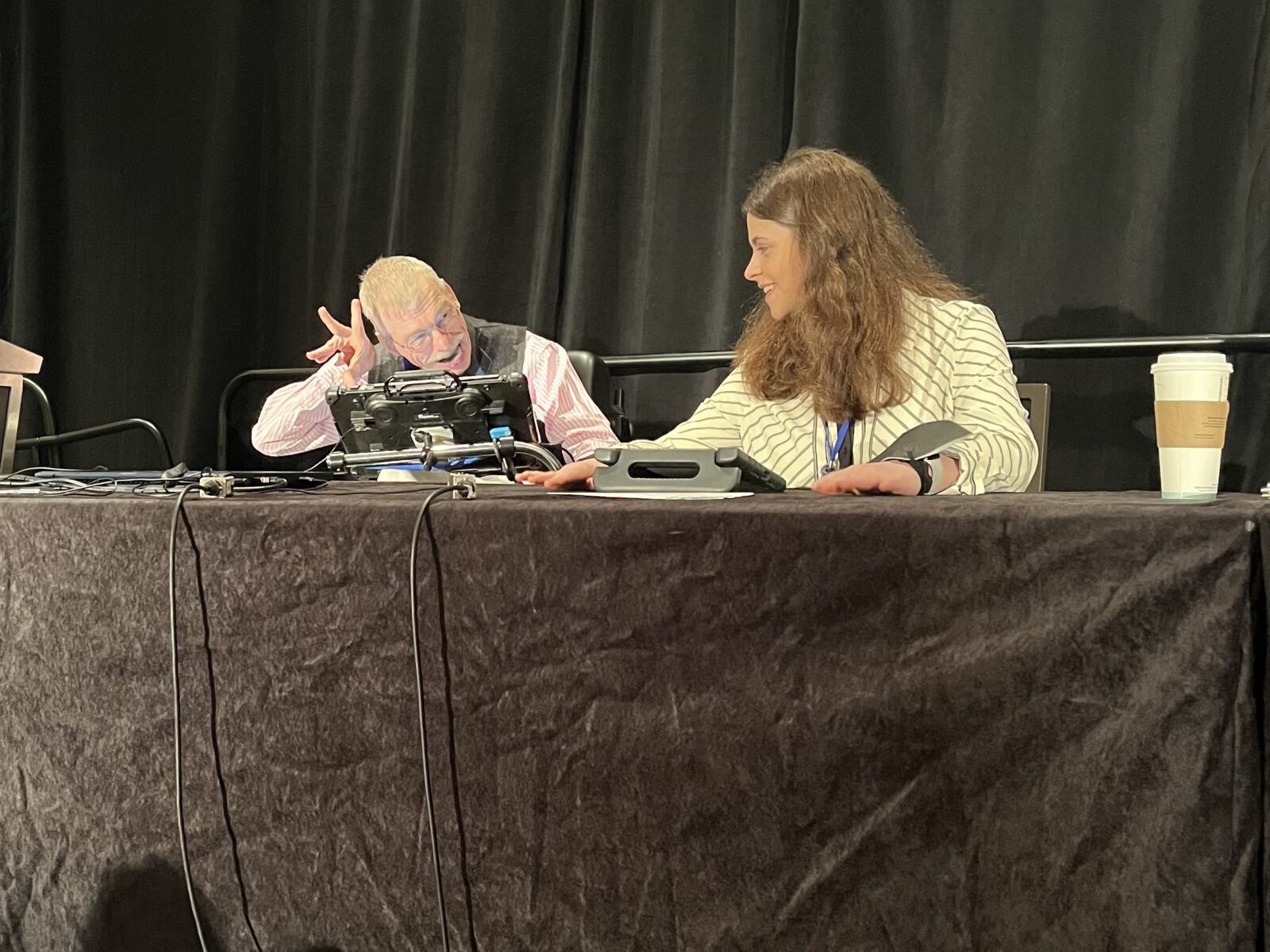 Bob Williams and Jordyn Zimmerman sit on a dais chatting with one another. They both have AAC devices in front of them.