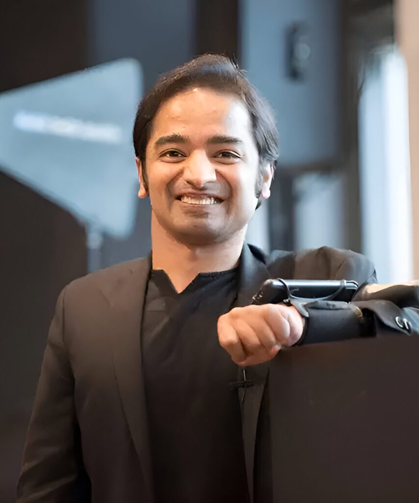 Godfrey Nazareth, a smiling man with brown skin who is looking at the camera and holding up his left arm to show his AAC device strapped on his forearm.