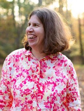 A white woman with shoulder length brown hair is standing outside with foliage and a sunset in the background. She is wearing a pink and white button down shirt. She is looking off to her right with a large smile.