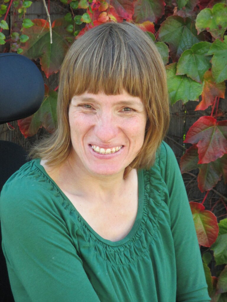 A white woman sitting in her wheelchair smiles broadly up at the camera. She has shoulder-length, straight, light brown hair and is sitting outside in front of some foliage.