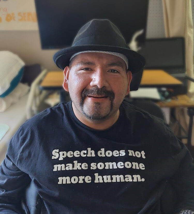 A Mexican-American man sits in a wheelchair in front of his bed and desk in a nursing home. He is smiling at the camera and wearing a hat over his bandaged head. He is wearing a black long-sleeved t-shirt that says "Speech does not make someone more human" in white letters.