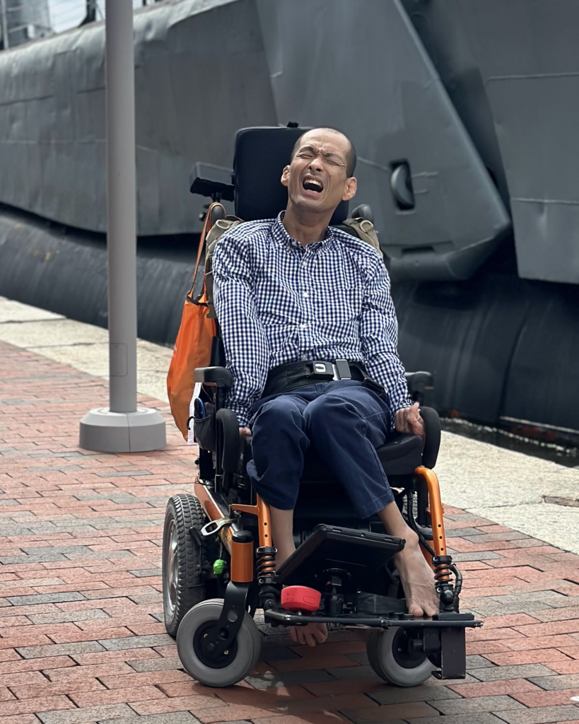 Tim Jin, a Korean-American man with a shaved head, sits in his power chair outside next to a docked naval vessel. He is smiling at the camera, wearing a long-sleeved blue and white checked shirt. His AAC device is positioned at his bare feet, with which he types.