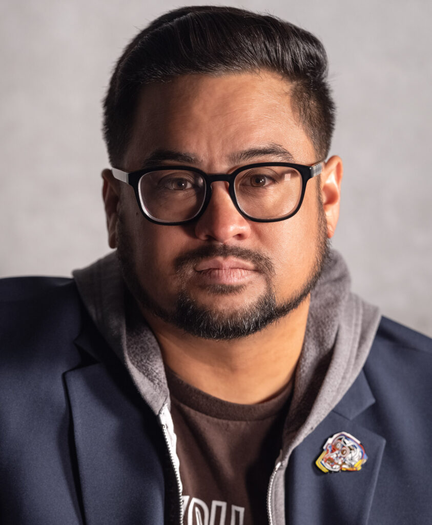 A Filippino-American man with dark brown hair, a goatee, and eyeglasses stares directly at the camera with a neutral, expressionless face. He is wearing a hoodie under a navy blazer.