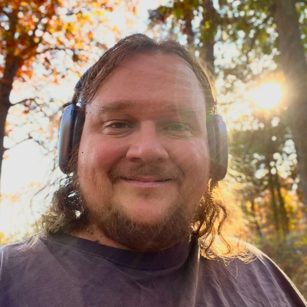 Chris Williams, a warmly smiling white man with long hair and facial hair, wears headphones while smiling down at the camera. He appears to be standing on the edge of a forest, with sunlight streaming through tall trees behind him. The sunlight hits right behind his head so that the left side of his head and hair is glowing.