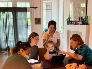 A smiling young girl uses her AAC device. Three women surrounding the girl cheer her on, and another woman sitting at the dining room table with them takes notes.