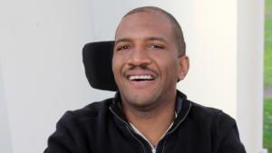 Lateef McLeod, a Black man wearing a black fleece top sits in his power wheelchair smiling at the camera.