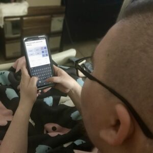 A glimpse over the shoulder of Ren, a fat nonbinary person with glasses and a partially shaved head visible, as they type into the AAC app on their smartphone. On their lap, a blanket is visible, patterned with smiling cartoon ghosts in the colors of the transgender flag.