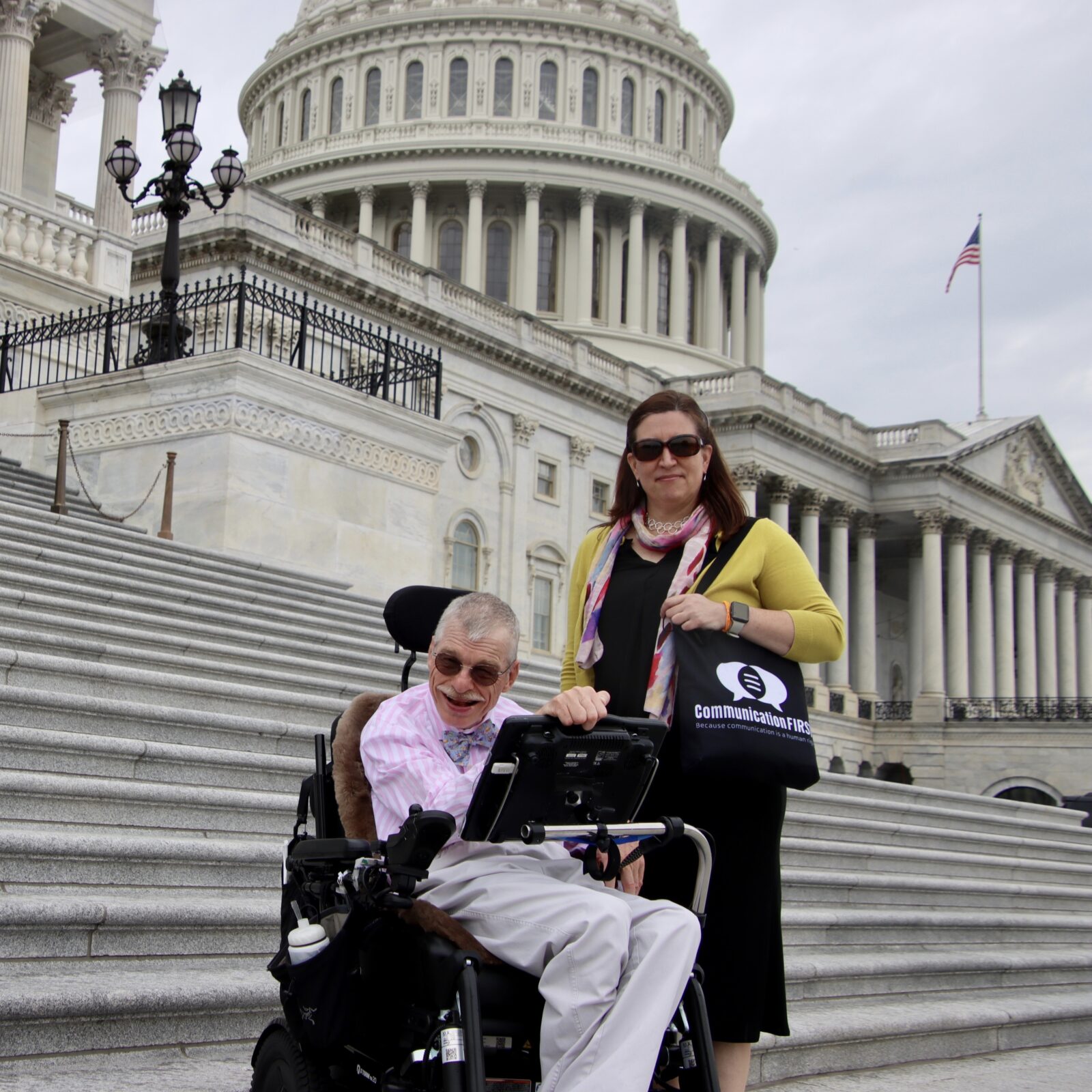 Outside the U.S. Capitol, CommunicationFIRST Policy Director Bob Williams poses with Executive & Legal Director Tauna Szymanski. The smiling co-founders sport sunglasses. An AAC device is fixed to Bob’s powerchair.