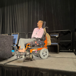 Tim Jin, a smiling AAC user with a shaved head, sits in his powerchair and presents onstage at the AAC Research Summit. Tim's AAC device is positioned by his bare feet, with which he types.