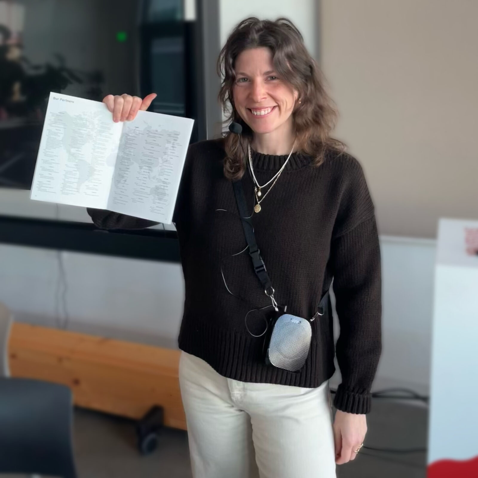 A voice amplification device hangs from a strap around Sophie Bohnen's shoulder, and its microphone attachment is positioned by her mouth. Sophie smiles and looks at the camera as she holds up a printed program.