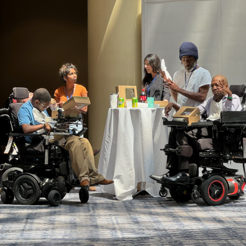 Alongside their aides, AAC users eat boxed lunches at a table in a hotel atrium.