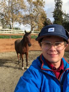 A masculine-presenting person in a blue hat, blue jacket, and gold glasses smiles while looking at the camera. Behind him, a brown, Arabian breed horse with a black mane and white socks stands relaxed, also looking at the camera.