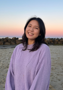 An East Asian young woman stands on a sandy beach, wearing a light purple sweater and looking at the camera. Behind her is a blue and pink sky.