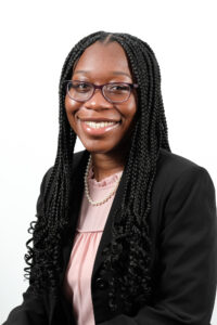 An African American woman with purple glasses and black knotless braids is smiling. She wears a pink blouse, a black blazer, and a string of pearls. Behind her is a white background.