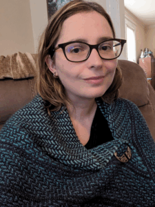 A fair-skinned person with light brown, shoulder-length hair, and dark brown eyes sits looking at the camera with a slight smile. She is wearing glasses and a knitted shawl fastened by a clasp.