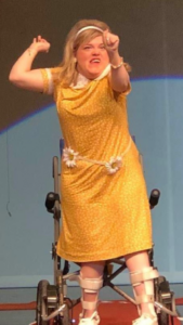 Marisa Conners stands in front of her wheelchair, fists raised triumphantly. She models a yellow floral dress she designed and a white headband.