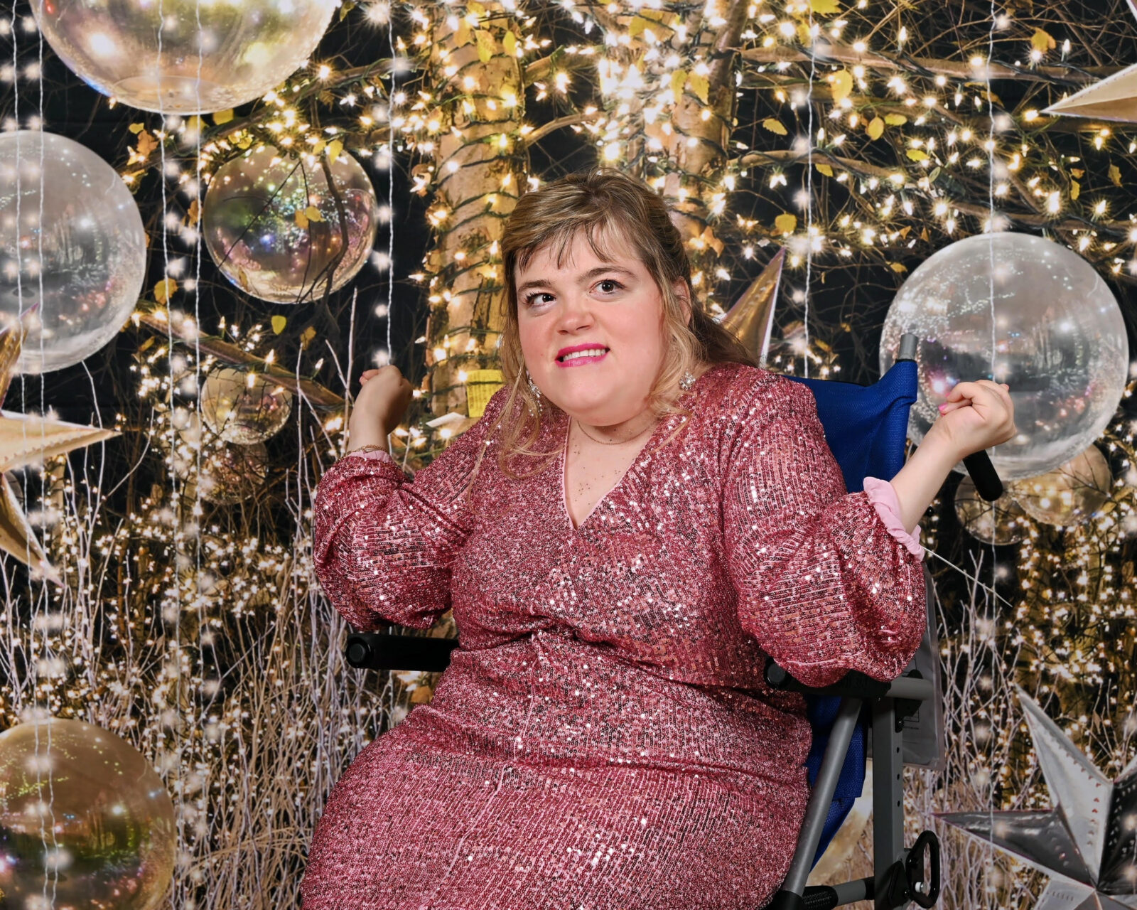 Marisa Conners wears a sparkling pink formal dress and poses in front of a background filled with golden lights, stars, and orbs.