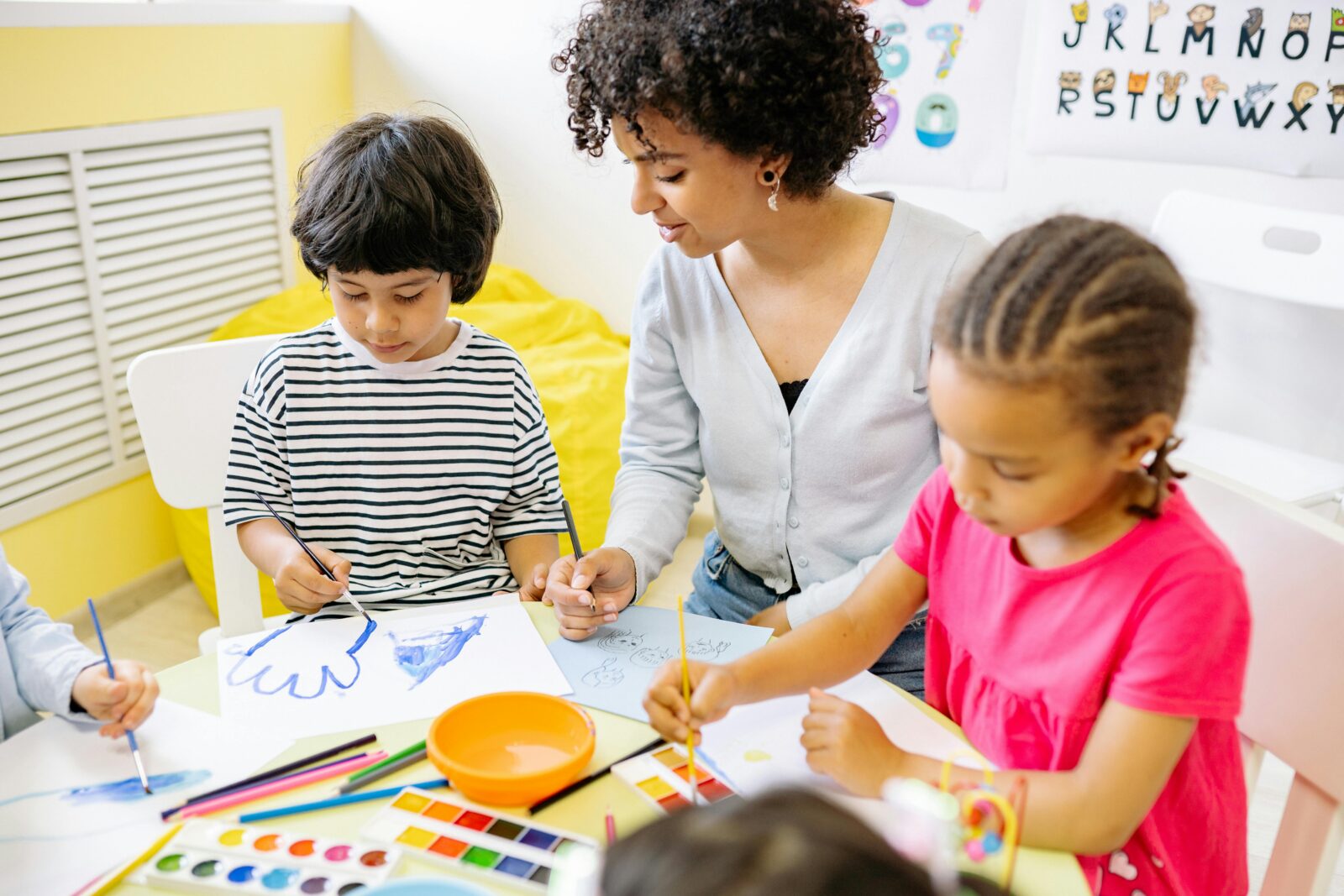 Young children sit at a circular school table and paint. A teacher sits with them, smiling at one of the paintings.