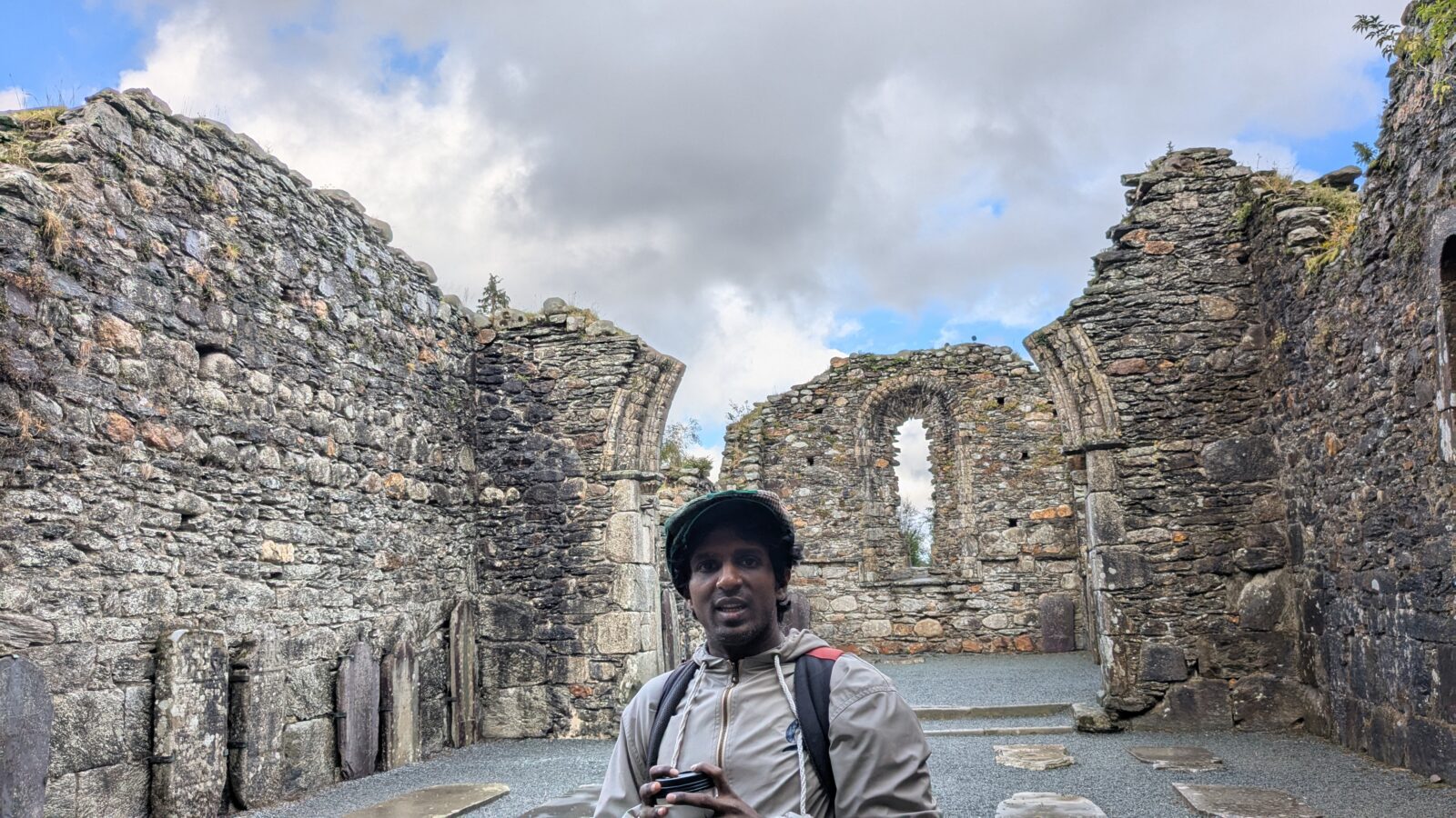 Wearing a dark cap and light brown jacket, Hari Srinivasan stands inside stone castle ruins along the coast of Northern Ireland.