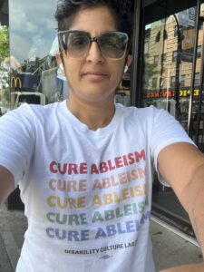 Shubha Balabaer takes a selfie while wearing glasses and "Cure Ableism" shirt.