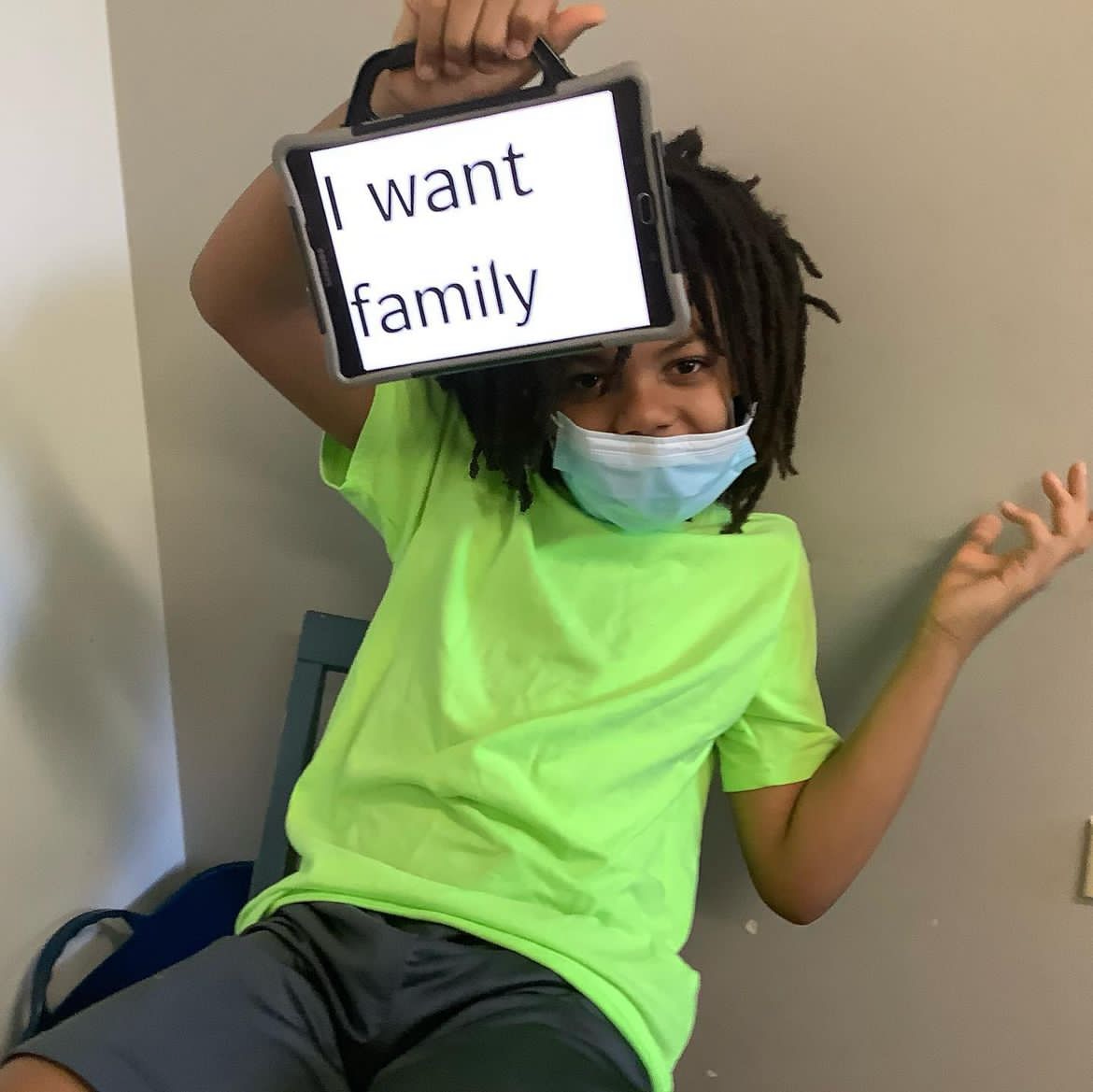 A young boy wearing a mask holds up a speech generating device with the sentence, "I want family."